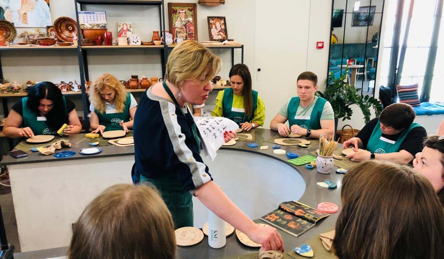 Instructor demonstrates a hands-on craft activity at a round table while participants work with clay and tools, representing community engagement and resilience supported through humanitarian relief efforts related to the war in Ukraine.