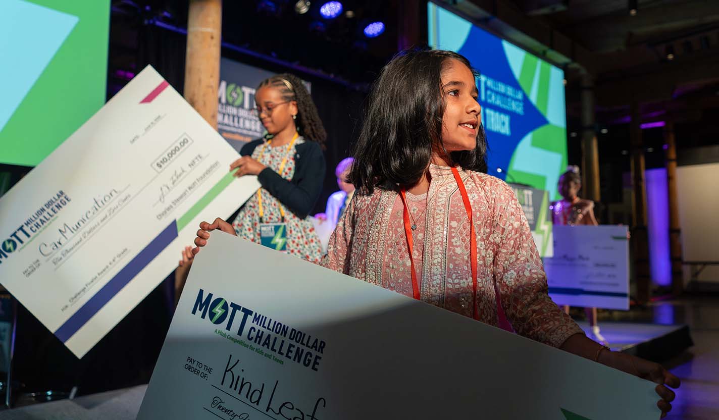 Young participants stand on stage holding large award checks during the Mott Million Dollar Challenge awards ceremony.