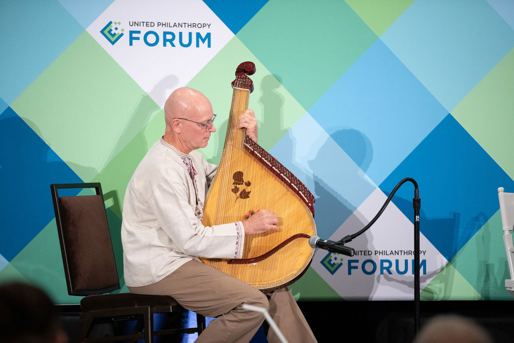 A seated man plays the bandura, a traditional Ukrainian plucked-string musical instrument, in front of a background with the United Philanthropy Forum logo.