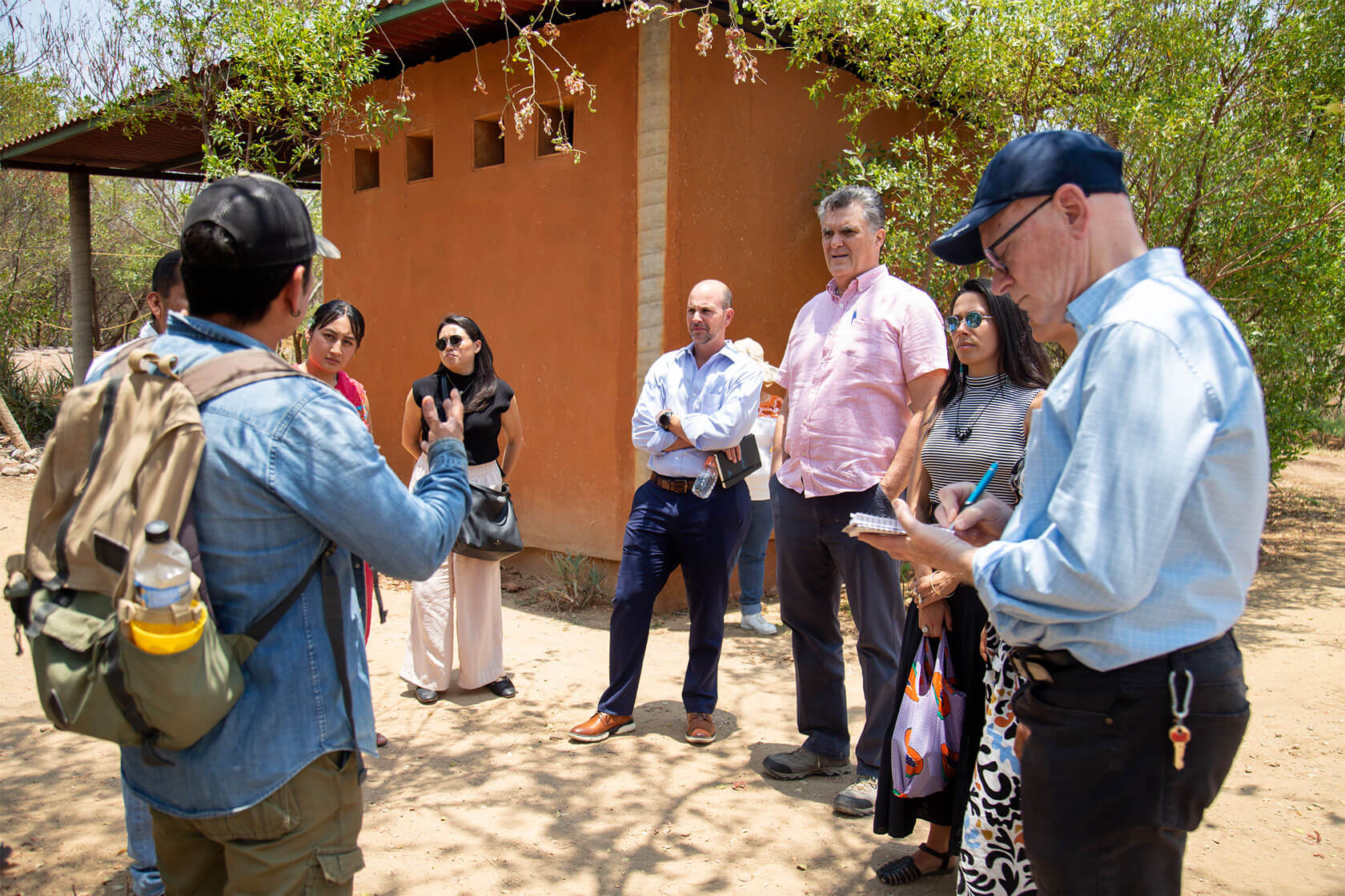 A small group standing outdoors on a sunny day listens intently to a speaker.
