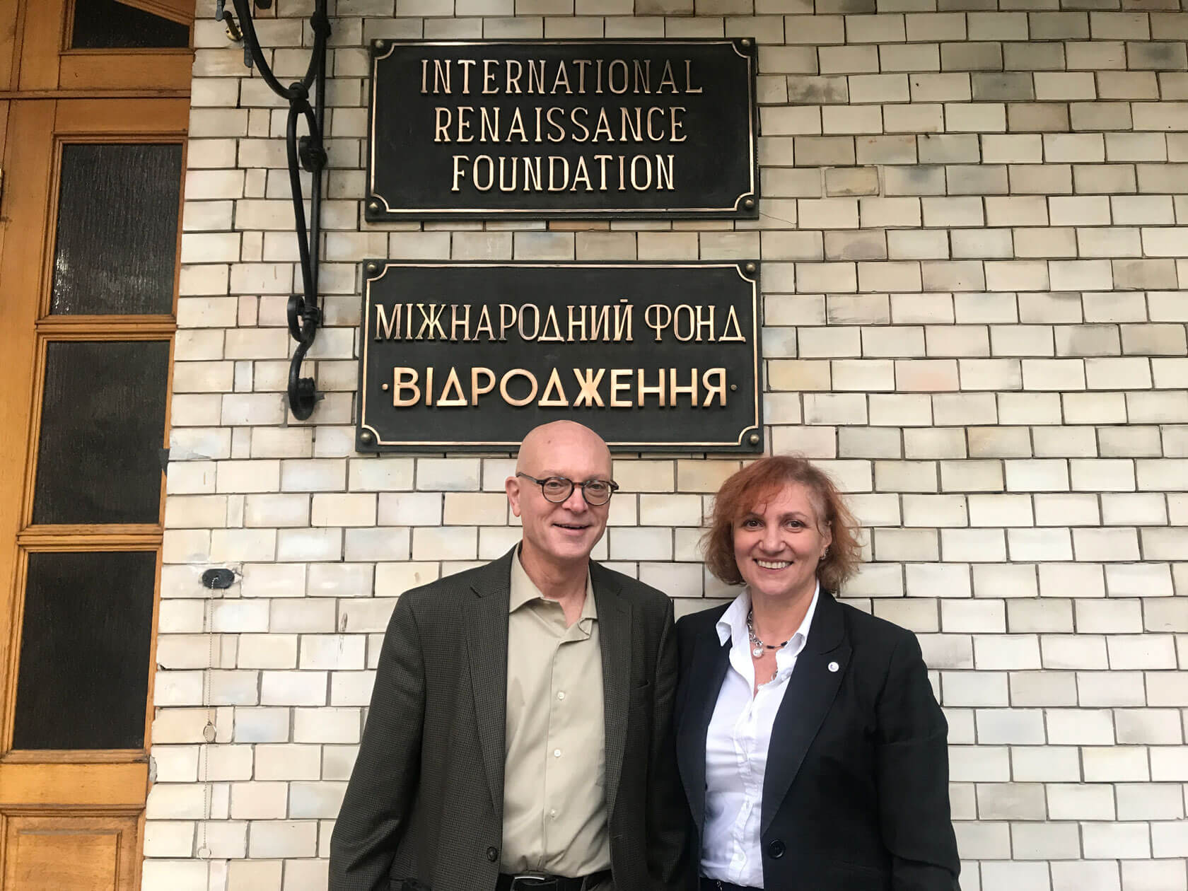 A smiling man and woman stand in front of a building beneath two black plaques mounted on the brick exterior. The upper plaque reads “International Renaissance Foundation,” and the lower plaque displays the organization’s name in Ukrainian.