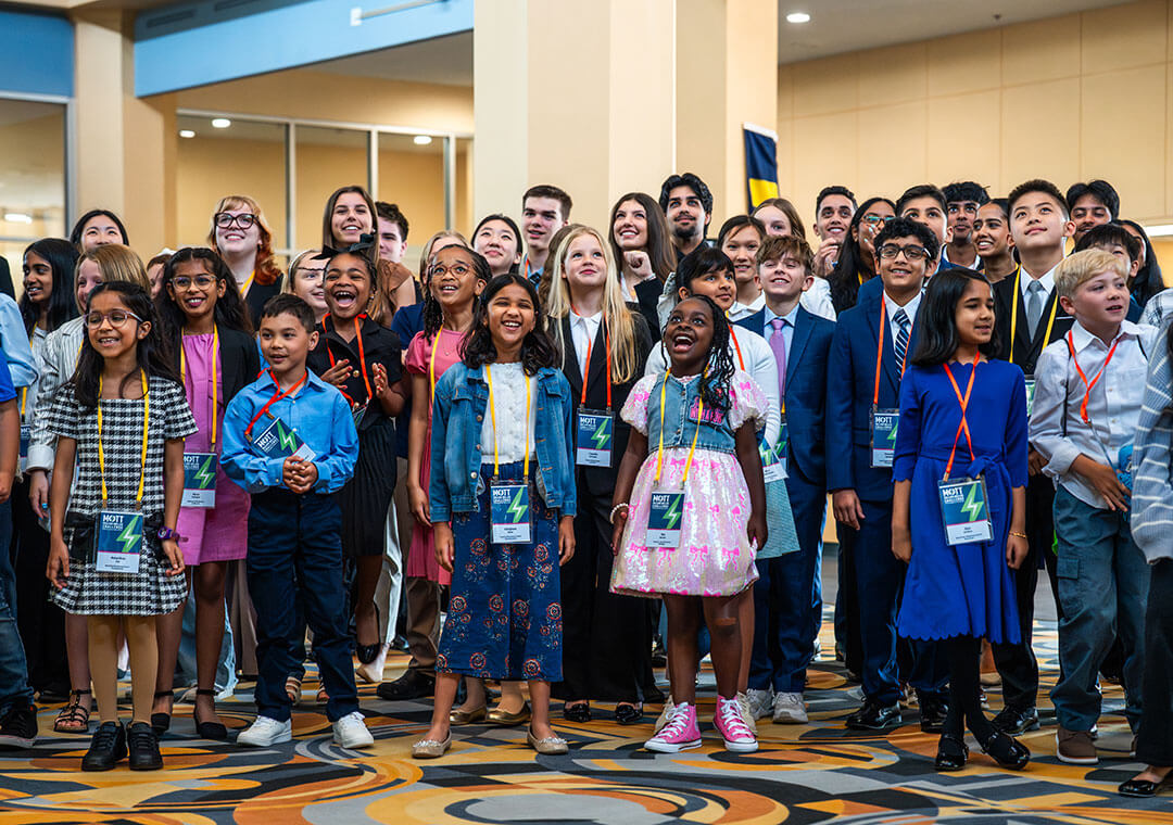 A large group of children and teens wearing event badges gathers for a photo, smiling and looking upward.