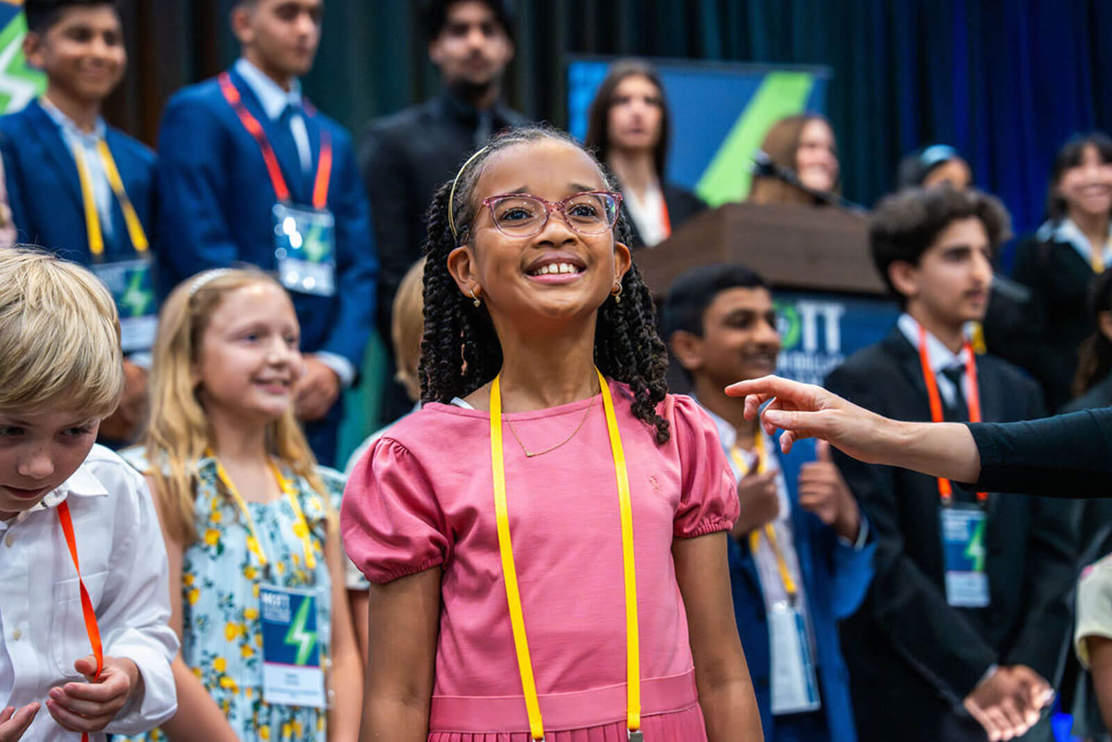 A young participant in a pink dress and glasses smiles while standing among other students onstage.