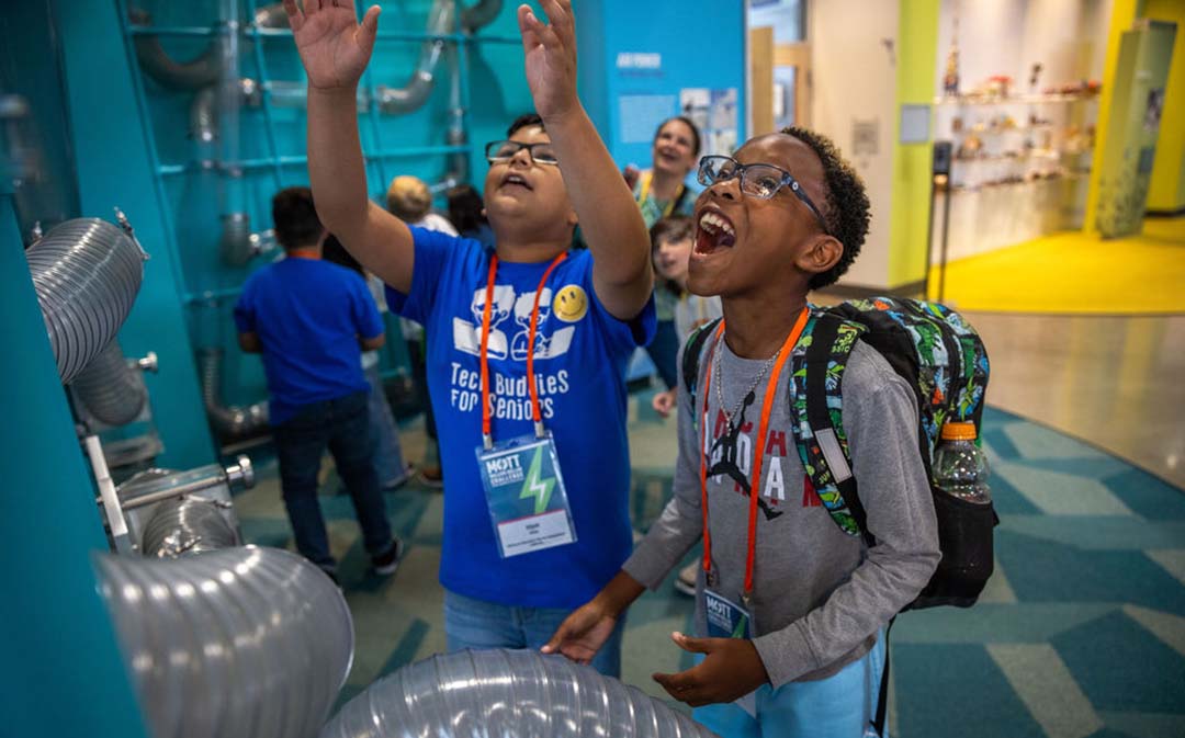 Two boys laugh and reach toward an interactive exhibit with large clear tubes.