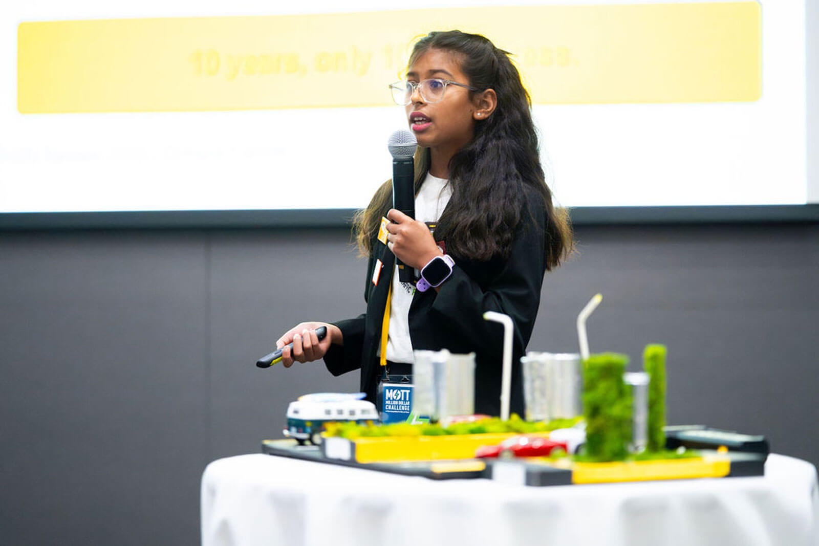 A young presenter speaks into a microphone beside a model featuring roads, buildings, vehicles and greenery.