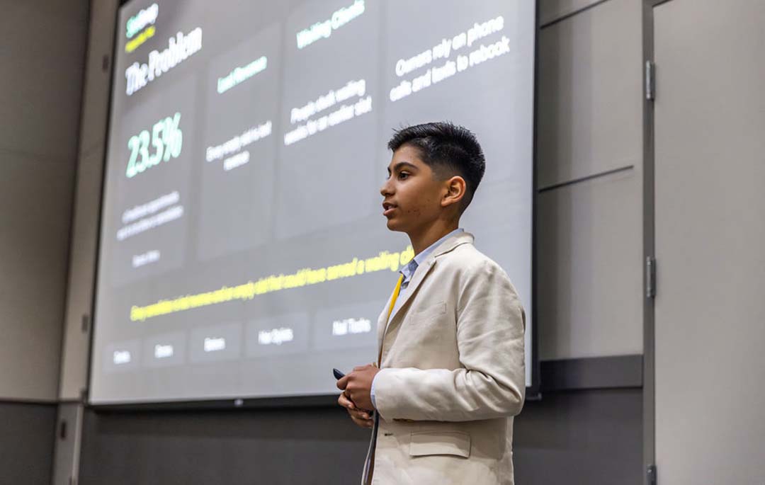 A young presenter in a light-colored suit stands before a projected slide showing research findings.