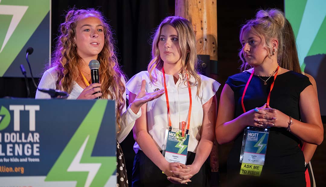 Three teenage students stand onstage as one speaks into a microphone and gestures with her hand.