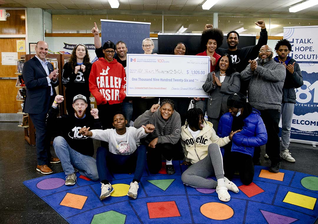 Students pose with a ceremonial Charles Stewart Mott Foundation check during a Youth Choice Awards presentation while another student photographs the moment inside a classroom.
