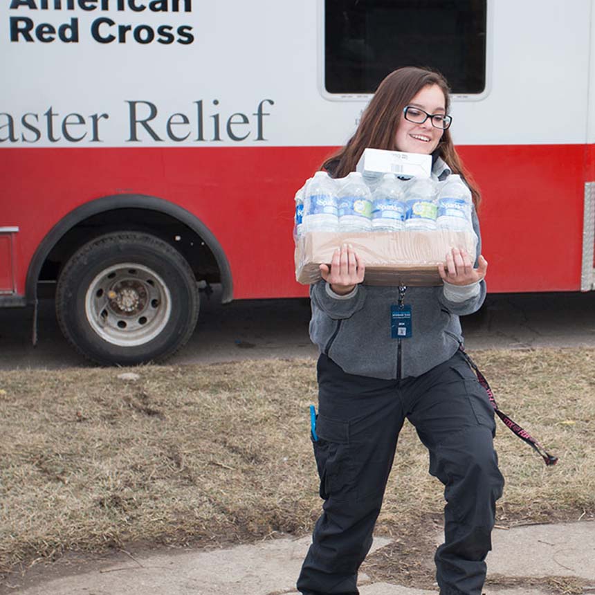 A smiling woman carries a flat of water bottles from an American Red Criss Disaster Relief truck.
