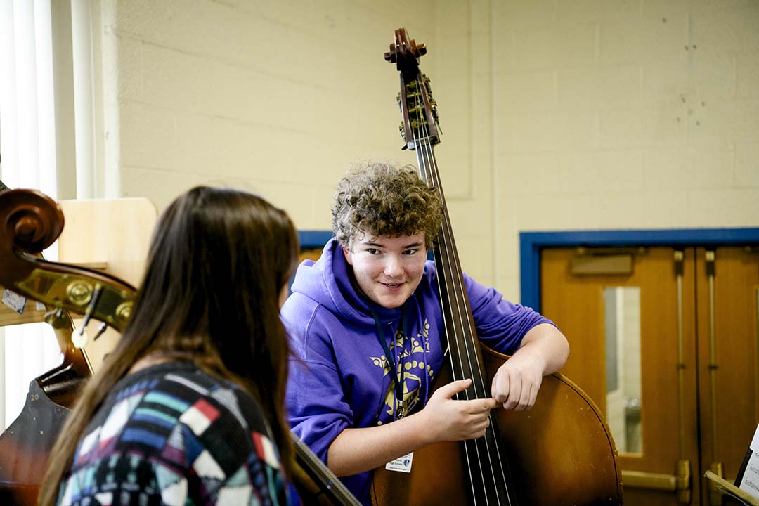 A teen smiles over his shoulder while leaning on an upright bass, with another musician seated in the foreground.