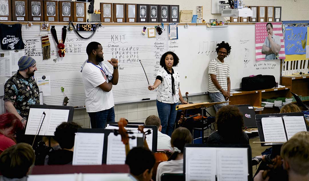 In an orchestra classroom, a student holding a bow addresses seated string players as three adults look on.