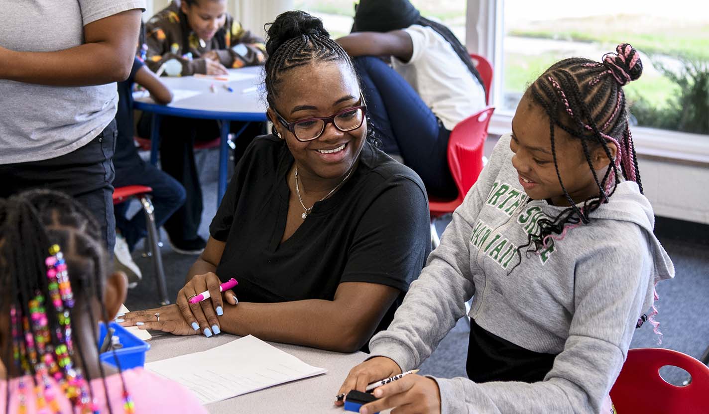 An adult works with children on a classroom activity while other children work at a table in the background.