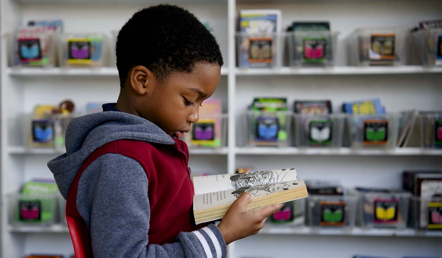 A child sits in a library reading an illustrated book, with shelves of colorful books in the background.