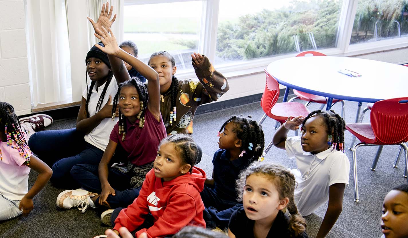 Children gather on the floor of a bright classroom, with several raising their hands to participate in a group activity.