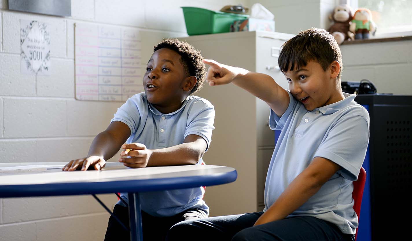 Two children in matching light blue shirts sit at a classroom table, with one extending an arm and pointing toward something outside the frame.