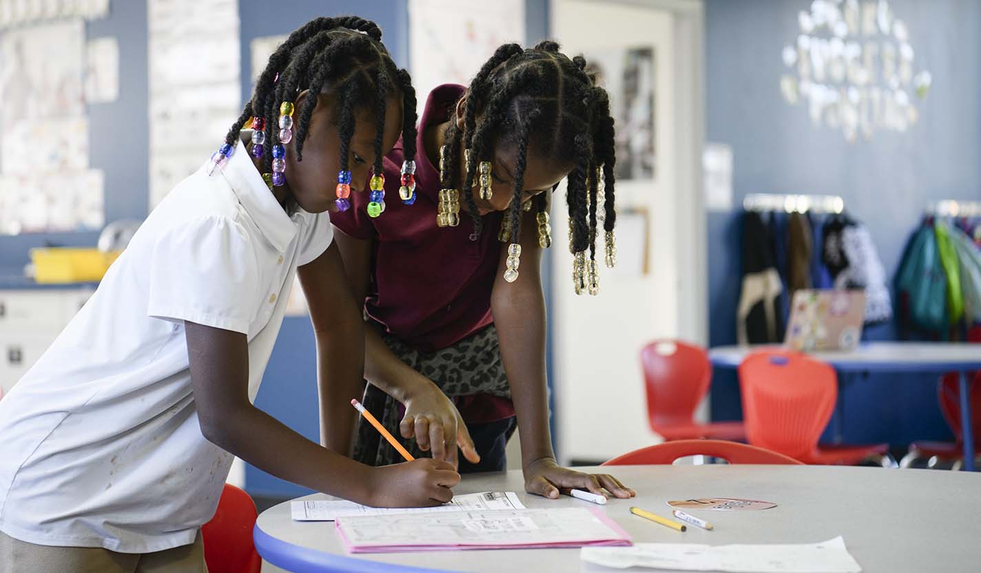 Two children lean over a classroom table and work together on a worksheet, with one writing in pencil and the other pointing to the page.
