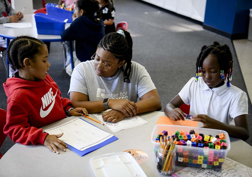 Two children complete hands-on learning activities with an adult at a classroom table using colorful counting cubes, pencils and worksheets.