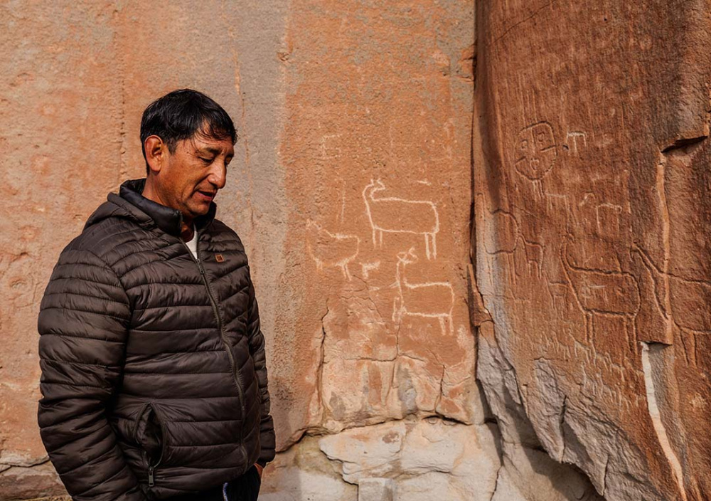 Person in a dark puffer jacket stands beside a weathered reddish rock wall featuring petroglyphs of etched figures, including several animals.