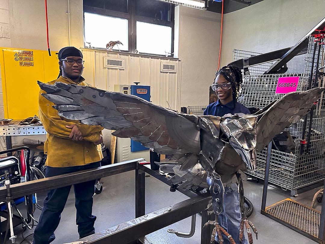 Two teens stand behind a welded metal sculpture of a bird.