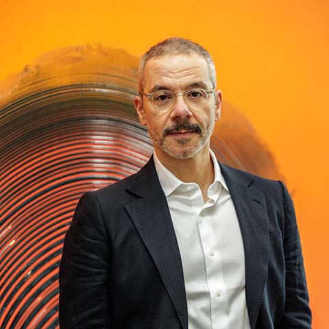 Person wearing glasses, a dark suit jacket and white shirt, standing in front of a vivid orange background with a large circular abstract artwork.