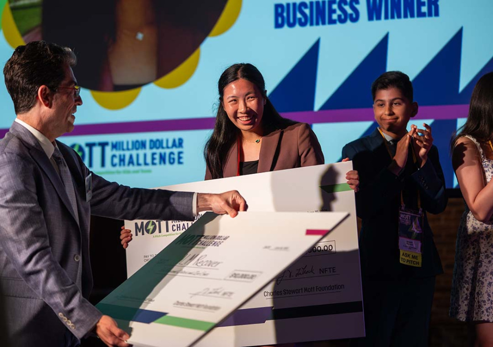 A group of young participants hold an oversized award check onstage during the Mott Million Dollar Challenge as an adult presenter stands beside them.